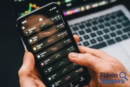 Pessoa usando um aplicativo de finanças no celular, com cotações e variações de ativos na tela. Ao fundo, um notebook com gráficos. A imagem representa o uso de simulador de investimentos. Logotipo Flávio Siqueira no canto inferior direito.