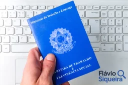 Pessoa segurando uma carteira de trabalho física azul sobre um teclado de computador branco, simbolizando a migração para a carteira de trabalho digital e a modernização dos registros profissionais no Brasil.