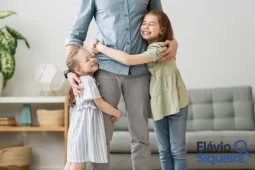 Pai em pé sendo abraçado por duas filhas sorridentes em um ambiente doméstico, representando um momento de carinho no dia dos pais.