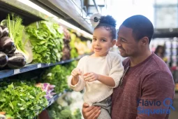 Pai segurando a filha no supermercado enquanto escolhem verduras, simbolizando compras da cesta básica.