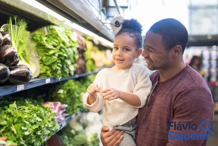 Pai segurando a filha no supermercado enquanto escolhem verduras, simbolizando compras da cesta básica.